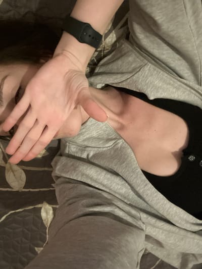 A person lies on a bed covered with a brown and gold patterned quilt. They are wearing a grey zip-up hoodie and a black top with a cutout. Their left arm, adorned with a black smartwatch, is raised, with their hand partially covering their face. Only one eye and a portion of their face are visible. The hoodie is partially zipped, revealing the neckline. The angle of the photo is slightly off-center.