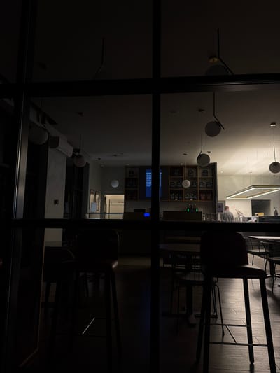 The image captures a modern interior space, likely a cafe or lounge, viewed through a dark window or glass partition at night. The photograph is oriented sideways, rotated approximately 90 degrees clockwise, causing the floor to appear as a vertical wall and the true walls to be horizontal.
Inside, the brightly lit area features contemporary design. Several spherical pendant lights are suspended at various heights, providing illumination alongside a large, glowing rectangular LED fixture on the ceiling. A lone person is seated at a high table or counter, facing away from the camera, seemingly focused on an item in front of them.
Against one wall is a wooden shelving unit with numerous cubbies, displaying various objects, possibly products or decor. Below this, a blue electronic screen with unreadable white text is visible. Further down the wall, an air conditioning unit is mounted, and a small red object, possibly a fire alarm or sensor, is affixed to a pillar.
The immediate foreground is dominated by dark, thick window frames or structural beams, forming a grid that partially obscures the interior view. Reflections of the interior lights and other elements are noticeable on the glass surface. The exterior beyond the window is completely dark, indicating the scene takes place at night. No specific weather conditions are discernible. The overall atmosphere appears quiet and modern. No discernible text is readable besides the unidentifiable white text on the blue screen.