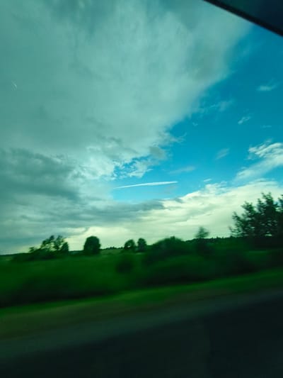 A blurred, horizontal, low-angle outdoor shot captures a vista of green fields and trees under a dramatic sky with clouds and patches of blue. The scene is taken from a moving vehicle, indicated by the motion blur. The sky is dominant, taking up the top two-thirds of the frame. It transitions from a teal-green hue on the left to a bright blue on the right. A faint white contrail is visible across the blue expanse. The lower portion of the image shows a line of trees and vegetation, also blurred by motion. The foreground consists of a dark, out-of-focus area, likely the road or the vehicle's interior. The overall impression is one of a fleeting glimpse of a landscape seen at speed. The image does not contain any recognizable landmarks or identifiable human activity.