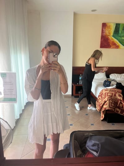The image displays an indoor scene within a hotel room in Chamilpa, Mexico. A female subject with dark hair pulled back, wearing gold hoop earrings, rings on both hands, a silver bracelet, a gold-toned bracelet, a black tank top, a white layered skirt, and a white sheer long-sleeved cover-up, is positioned centrally, taking a mirror selfie with a light-colored smartphone.

In the background, to the right, a second female subject with long blonde hair, dressed in a black sleeveless top, black cargo pants, and white Converse-style sneakers, stands next to a bed. She is holding an orange bottle or container in her right hand.

The room features light yellow-cream painted upper walls transitioning to white lower walls, and light brown square floor tiles with darker grout. The bed has a dark brown wooden headboard, white linens, and a brown and orange geometric patterned blanket or throw. A blue bag is also on the bed. A dark brown wooden nightstand sits beside the bed, holding a clear water bottle. Above the bed, a two-part abstract painting displays shades of red, orange, yellow, and green. On the left side of the room, white sheer curtains cover a window. A white portable fan with a circular grille is visible on the floor near the mirror, alongside a white sign with green text, possibly a hotel notice. An open black suitcase with inner compartments and contents is positioned in the immediate foreground, at the bottom of the mirror frame.