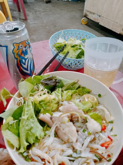 A close-up, high-angle shot shows a meal with a can of Tiger beer and a plastic mug of what appears to be beer on a red table in what appears to be a public space. The main focus is a white bowl filled with Vietnamese noodles (likely Banh Canh) and vegetables. The noodles are submerged in broth and topped with sliced meat and leafy greens. A smaller blue basket of chopped lettuce and a pair of black chopsticks sit nearby. The can of Tiger beer is placed to the left of the bowl. The background shows a concrete floor and a partially visible cardboard box on wheels, suggesting the setting might be a street-side eatery in Ea Drăng, Vietnam.