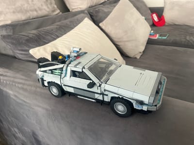 A detailed, forensic, matter-of-fact description of the scene is provided below.
A model of the DeLorean time machine from the movie "Back to the Future" is displayed on a gray sectional sofa. The model is constructed from light gray and white Lego bricks, with some black, blue, and transparent elements. It features detailed components such as wheels, headlights, taillights, a flux capacitor, and other elements seen on the movie car. The sofa is upholstered in a dark gray fabric, and several throw pillows of various shades of gray and off-white are arranged behind the model. In the background, on the sofa, are a red hat, a small blue object, a red notepad, and a smartphone with a light blue case. The lighting suggests an indoor environment, possibly a living room. The overall scene is static, with no people or actions depicted. The location is not specified in the image.