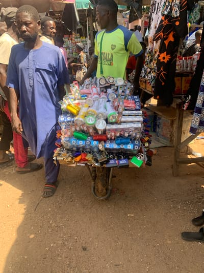 This media file captures a vibrant street market scene in Boi A, Nigeria. The primary subjects are several men, some of whom appear to be vendors or customers. One man in the foreground wears a blue pinstriped garment and red trousers. To his left, another man in a yellow and blue shirt is partially visible. Behind them, a stall is laden with a variety of goods, including numerous small boxes, packaged toothbrushes or similar items, various colorful brushes, and an assortment of padlocks in different colors. The items are arranged on a cart, suggesting a mobile vendor.

The scene is outdoors, with the ground covered in dirt and small stones, typical of a rural or semi-urban market environment. Sunlight casts distinct shadows, indicating it is likely daytime, possibly late morning or early afternoon. The overall atmosphere seems busy, with the arrangement of goods and the presence of multiple people suggesting commerce is actively taking place.

Visible text includes brand names like "Xtreme," "Olive," and "STAR FORCE XFORCE," as well as the partial word "SALA" and "GIOUP" on the yellow shirt. These details point to the types of products being sold, such as personal care items and hardware. The image is taken from a slightly elevated, tilted perspective, offering a dynamic view of the market stall and its surroundings.