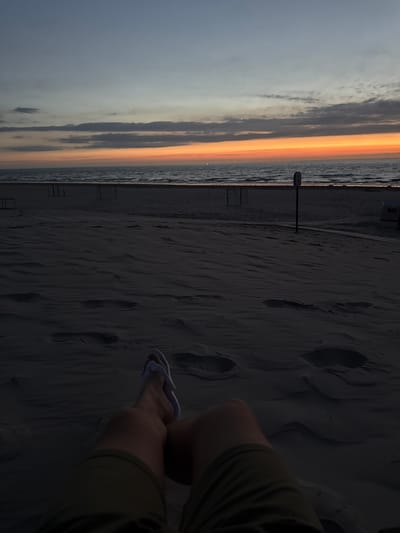 A person's legs and feet are visible in the foreground, with the person wearing green shorts and white flip-flops. They are sitting on a sandy beach in Langeoog, Germany, facing the ocean. The sun is setting, casting an orange glow on the horizon and the clouds. The sky is a gradient of blue and orange. In the background, there are some beach chairs and a soccer net. The overall scene is peaceful and serene, capturing the beauty of a sunset at the beach.