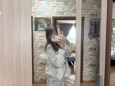 A young woman with long brown hair pulled into two buns is taking a mirror selfie in a room. She is wearing a white long-sleeved sweatshirt with white fringe detailing on the sleeves and light-wash jeans. In her right hand, she holds a white smartphone, capturing the reflection of herself and the surrounding environment.

The background features patterned wallpaper with a brick-like design and small images of what appear to be Venetian canals. To the left of the woman, on the wall, is a framed artwork of a leopard. To the right, reflected in the mirror, is a rack displaying several medals, suggesting athletic achievements. A doorway is visible, leading to another room where a bed can be seen. The floor is covered with wooden laminate flooring. The room appears to be a bedroom or dressing area. The image is taken indoors.