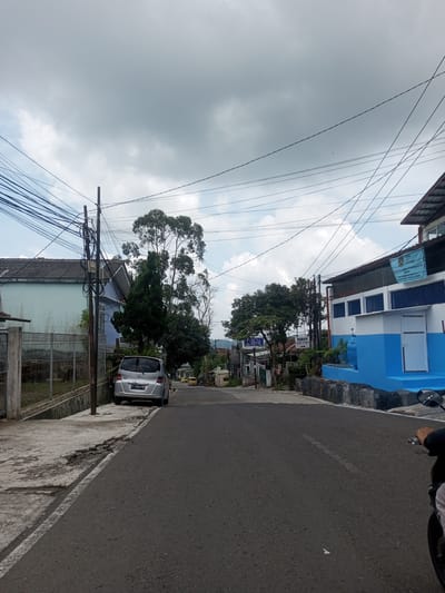 The image captures a street scene, focusing on a road in a residential or semi-rural area. The sky is overcast, with dense clouds obscuring much of the sky. Power lines are visible crisscrossing above the road.
On the left side of the road, there is a house with a blue-painted exterior and a fence. A silver car is parked along the roadside. The road itself is asphalt.
To the right, there is a building with a blue and white facade. A sign on the building reads "BADAN GIZI NASIONAL" and includes text "SATUAN PELAYANAN PEMERIKSAAN GIZI (SPPG)". Additionally, a sign on the left of the blue and white building reads "SETIA HUT".
A motorcycle is visible on the right, with the rider's arm and part of the motorcycle visible in the bottom right corner of the frame. The location in the city of Ban Phouviengnoy, Laos is not evident in the image.