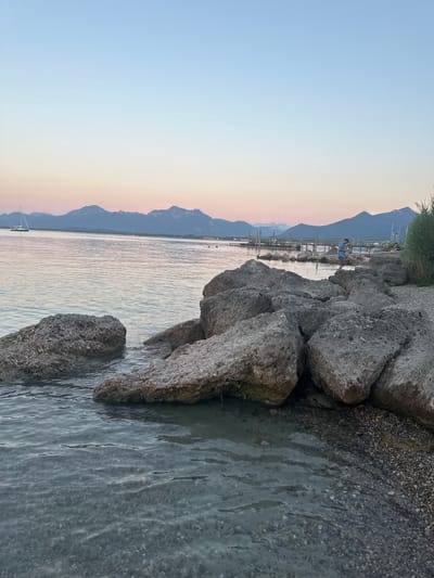 The image depicts a tranquil lakeside scene at dusk, likely in Prien am Chiemsee, Germany. The sky is a gradient of soft pinks and blues, with distant mountains silhouetted against the horizon. A calm body of water, possibly Lake Chiemsee, occupies the majority of the frame, reflecting the subtle colors of the sky. Large, rough-textured rocks are scattered along the shoreline, with smaller pebbles and gravel visible where the water meets the land.
In the middle ground, a wooden pier extends out into the lake. A person wearing a blue shirt and shorts is standing on the pier, facing away from the camera, seemingly engaged with something on the pier or in the water. Another person, partially obscured by vegetation, sits on the pier to the right. A sailboat with its mast visible is anchored in the water to the left, and another small boat can be seen further out. Lush green vegetation lines the far right edge of the image. The water is clear enough to see the gravelly bottom near the shore.
