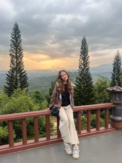 A young adult female is positioned centrally, seated on a dark brown wooden railing with her legs crossed at the ankles. She faces the viewer with a slight smile, her long, wavy brown hair draped over her shoulders. She wears a brown hooded jacket over a black top, beige wide-leg cargo pants, and light-colored chunky-soled sneakers. A black crossbody bag hangs from her left shoulder. Her hands rest on the top bar of the railing, which is supported by vertical balusters. The ground beneath the railing is a dark grey tiled or paved surface. To the far right, a traditional East Asian style multi-tiered stone lantern or decorative pillar is situated on the railing.
The background presents a panoramic view of a mountainous and hilly landscape located in Thôn 8, Vietnam. Two tall, slender, conical evergreen trees frame the female subject. Dense green vegetation covers the undulating terrain, and in the mid-distance, a valley contains scattered buildings and structures consistent with a town or settlement. The sky is heavily overcast with dark grey clouds, but a distinct horizontal band of warm orange-yellow light is visible along the horizon, suggesting either a sunrise, sunset, or sunlight breaking through the cloud cover.