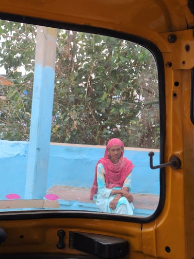 An elderly woman, wearing a pink headscarf and a light blue and pink outfit, sits on a ledge. Her arms are crossed in front of her. She is viewed through the passenger side window of a yellow auto-rickshaw, with the yellow interior of the auto-rickshaw framing the lower part of the image. The woman is seated in front of a bright blue wall, with green foliage visible in the background. The scene appears to be in Jodhpur, India, based on the auto-rickshaw and the style of dress.