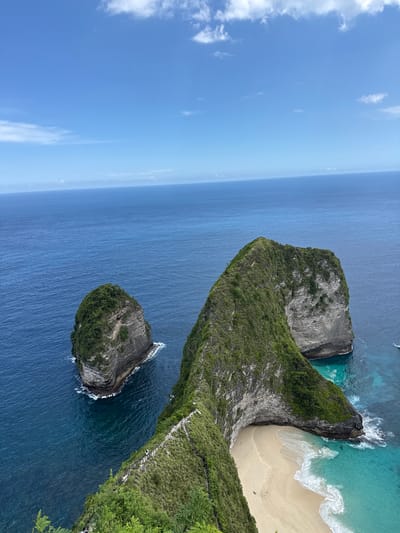The image shows an aerial view of Kelingking Beach in Bunga Mekar, Nusa Penida, Indonesia. A large, T-Rex shaped rock formation dominates the scene, covered in green vegetation. A sandy beach is nestled at the base of the formation, bordered by turquoise water and white waves. To the left, the deep blue ocean stretches to the horizon, meeting a pale blue sky with scattered white clouds. A smaller, rocky island, also covered in vegetation, is located in the water. A visible pathway with people is seen on the top-right part of the image.
