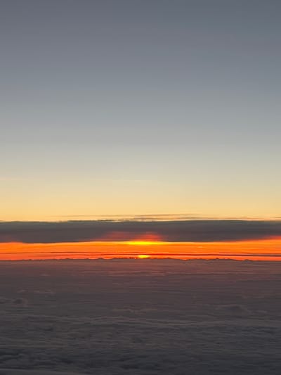 The image depicts a sunrise or sunset view from above the clouds. The sky transitions from a dark gray on the right to orange and then to a lighter gray on the left. The sun is obscured by clouds but the bright orange colors suggest it is near the horizon. The scene appears to be taken from an airplane, the clouds below appear as a flat, textured surface. The location is unknown and there is no information to confirm it is in Orp-Jauche, Belgium.