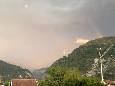 The image depicts a mountainous landscape, likely taken from a vantage point within or near the city of Cluses, France, given the context provided by the hints. The foreground shows the rooftops of houses with red tiles, suggesting a residential area. A prominent feature in the mid-ground is a dense, green forest covering the slopes of the mountains. To the right of the image, a tall yellow and silver construction crane stands against the mountainside, indicating ongoing development or maintenance work. A faint rainbow arcs across the sky above the forest-covered hills, partially obscured by hazy, reddish-orange clouds that create an atmospheric effect. The sky transitions from a lighter, hazy tone on the left to darker, more ominous clouds on the right, where the rainbow is visible. In the distance, nestled within the valleys between the mountains, appear to be scattered buildings, suggesting the presence of a town or village. The overall scene conveys a sense of natural beauty contrasted with human activity, possibly under unusual atmospheric conditions.