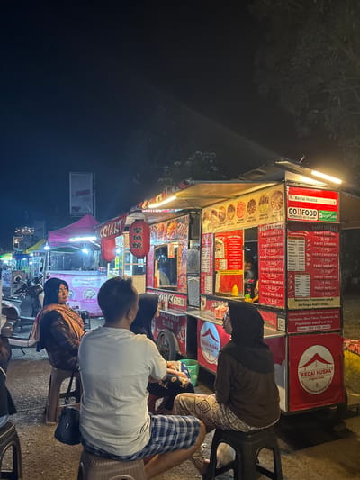 The image depicts a nighttime scene at a street food stall in Tanggul, Indonesia. A food cart, labeled "Kedai Husna," is brightly illuminated, displaying a menu with various food and drink packages. Several people are seated on stools, appearing to be customers or patrons of the stall. One woman in the foreground is wearing a headscarf and traditional clothing, while a man next to her is in casual attire. Another woman in a black headscarf is seated further back. The environment suggests a bustling street market with other stalls visible in the background, indicated by tents and lights. The overall atmosphere is lively, typical of a night market in Indonesia.