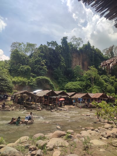 In Pantonrayeuk Satu, Indonesia, a scene unfolds by a river with a rocky shore. A row of open-air bamboo and thatched-roof huts lines the riverbank, some with people seated inside or standing nearby. In the water, several individuals are present. On a large rock in the river, a young woman in jeans and a light-colored top sits, while two other young women, one in a green swimsuit and another partially submerged, interact with her. Further downstream, a few more people are visible in the water. The river flows over rocks and around submerged stones. The background features a steep, vegetated cliff face covered in lush green trees and foliage. The sky above is a mix of blue with scattered white clouds. The overall atmosphere suggests a recreational area by a river in a tropical, natural setting.