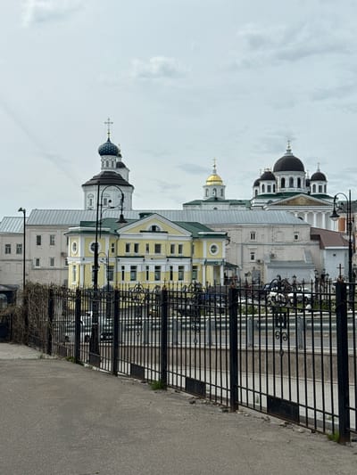 The image shows a vertical view of buildings in Arzamas, Russia. A black iron fence runs along the right side of the image, beside a gray asphalt road. Beyond the fence are several buildings, including a yellow and white building with a green roof and a white building with multiple black domes. The sky is overcast.