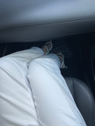 The image shows a close-up view of a person's legs and feet inside a vehicle, likely a car. The person is wearing white pants and open-toed sandals adorned with rhinestones. The sandals are white with silver straps that wrap around the ankles. One foot is resting on what appears to be a car's floor mat, which has a textured, grid-like pattern. The background consists of the car's interior, with dark gray leather seats and trim visible. The overall lighting suggests it is daytime, but the interior of the car is somewhat dim. The image is taken from a low angle, focusing on the lower half of the body and the immediate surroundings within the vehicle. There is no visible text in the image. The context of Coșernița, Moldova is provided, but there are no specific visual cues in the image to confirm this location.