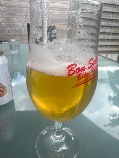 A partially filled glass of golden beer with a foamy head sits on a glass table outdoors. The beer glass has red lettering on it that says "Bon Secours Bière". To the left of the beer glass is a white can with red lettering that appears to be a Stella Artois beer can. In the background, a wooden fence can be seen. The scene appears to be from a patio or garden area, likely in Ath, Belgium given the "Bon Secours Bière" branding, as Bon Secours is a brewery located in Ath. The lighting suggests it is daytime.