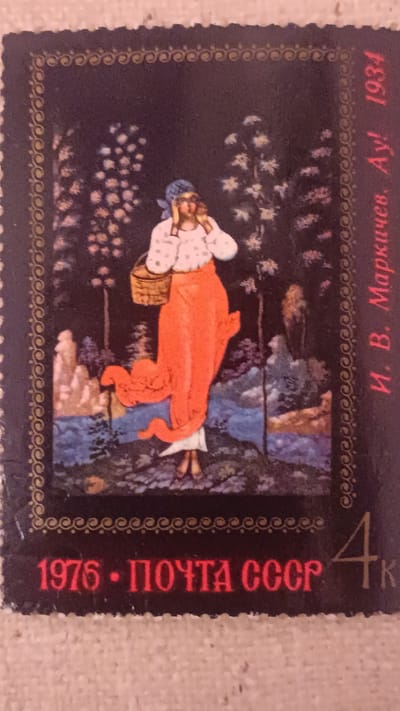 This is a close-up image of a postage stamp with a decorative border. The central image on the stamp depicts a woman in traditional attire with a blue headscarf and a long, flowing orange skirt. She is carrying a woven basket and appears to be walking through a wooded area with a body of water. The background features trees and rocky formations. Text in Cyrillic script is visible on the right side of the stamp, which reads "И. В. Маркичев. Ау! 1934". At the bottom of the stamp, the text "1976 · ПОЧТА СССР" is also visible, along with the numeral "4K". The image does not contain any recognizable landmarks or details that would indicate a specific location in the city of Onaldy, Turkmenistan.