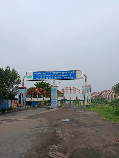 The image shows the Land Ports Authority of India border crossing at Benapole, Bangladesh. A large blue sign over the entrance reads "WELCOME TO THE REPUBLIC OF INDIA" in English and other languages. The gate is green with red detailing, and a fence runs along the sides. On the pillars of the gate, there are murals of historical figures, including one resembling Mahatma Gandhi. The road leading to the gate is wet and appears to have puddles, suggesting recent rain. Green grass and trees are visible on the sides, and in the background, a large building with a curved roof can be seen. The sky is overcast.