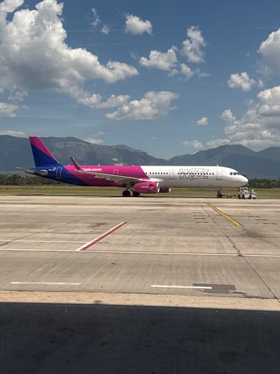 An airplane with the Wizz Air logo is parked on a tarmac at the Tirana International Airport, located in the vicinity of Kruja, Albania. The aircraft is predominantly white with a pink and blue tail section. It is being guided by a tow tractor. The airport tarmac is marked with yellow and white lines, indicating taxiways and runways. In the background, mountainous terrain is visible under a blue sky dotted with white clouds. The image is captured from a slightly elevated perspective, possibly from within the airport terminal.
