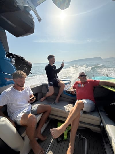Three adult males are observed on a moving motorboat on a large body of water, identifiable as the Sea of Galilee near Migdal, Israel. The scene occurs on a sunny day with a clear blue sky, indicated by the high sun causing a lens flare at the top of the frame.
The boat is a modern motorboat with blue and grey seating and decking, actively generating a wake. On the left, a male with curly light brown hair, wearing a white t-shirt and grey shorts, is seated and looking down at a smartphone in his hands. In the middle, a male with short light brown hair, dressed in a black long-sleeved athletic shirt and black patterned shorts, is seated and holding a black smartphone aloft, appearing to capture media of the distant landscape. On the right, an older male with short hair, wearing sunglasses, a red t-shirt, and light grey shorts, is seated with his legs crossed, holding a clear cup with a reddish liquid. All three individuals are barefoot.
Two wakeboards or surfboards, one yellow-green and one light blue, are stored along the right side of the boat. A black bag or watersports equipment is visible near the blue hull on the left, and a neon green life vest is partially visible on the deck. Hazy landmasses or mountains are visible on the horizon across the water.