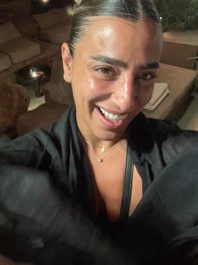 A forensic description of the image reveals a woman captured in a selfie at night within an outdoor lounge environment in Psarou, Greece. The woman, appearing in a medium close-up, has a tanned complexion, dark hair slicked back, and a broad smile, exposing white teeth. She wears a dark-colored, likely black, open-front top. Visible accessories include two delicate gold necklaces, one with a hamsa hand pendant and another with a round, clear stone pendant. She has stud earrings in her earlobes, a helix piercing in her right ear, and a piercing on her right nostril. A black strap, consistent with a handbag, is visible across her chest. The blurred background features low-set, natural-toned lounge furniture, possibly stone or concrete structures topped with light-colored cushions. A low table in the mid-ground holds a dark, decorative object containing a lit candle or lantern, providing warm ambient illumination. The overall setting suggests a dimly lit, upscale outdoor relaxation area.