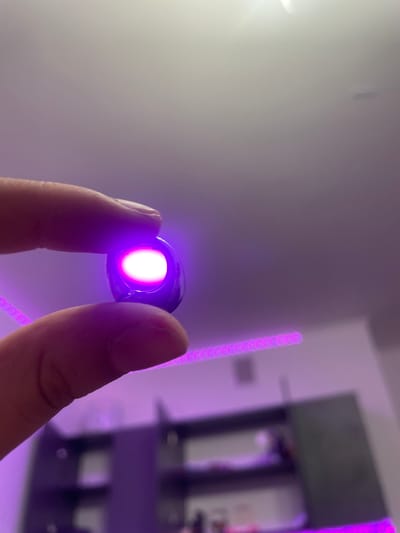 A close-up shot captures a person's fingers holding a small, dark, oval-shaped object that emits a bright pink light from its center. The background is softly lit and blurred, with hints of furniture and what appears to be a purple LED light strip running horizontally across the scene. The overall impression is of a dimly lit room with modern lighting elements. The image does not contain any discernible information related to the city of Romanovka, Russia.