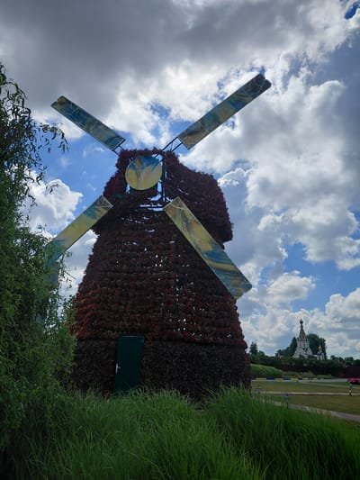 In Kovalivka, Ukraine, a decorative windmill structure covered in red and dark foliage is the central subject. The windmill has four large, rectangular blades with a painted abstract design of blue, yellow, and white. A dark green door is visible on the front of the windmill's base. In the background, a white domed church with a golden spire is set against a sky filled with dramatic clouds. The foreground is dominated by tall, green grass, and to the left, a weeping willow tree hangs over the scene. The overall environment appears to be a well-maintained park or garden.