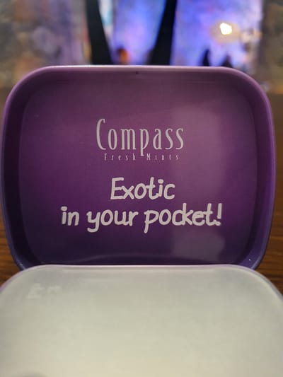 The image shows the interior of an open purple tin. The words "COMPASS Fresh Mints" are printed in white script at the top of the tin. Below this, also in white script, are the words "Exotic in your pocket!". The tin is sitting on a wooden surface. The background is blurred and shows colorful lights. The location in Malia Municipal Unit, Greece, is not discernible from the image.