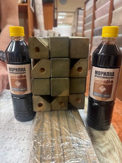 In this image, two bottles of "MORILKA PROFESSIONAL" wood stain, labeled in Russian as "МОРИЛКА ПРОФЕССИОНАЛЬНАЯ" and indicating a volume of 0.5L, are placed on top of a stack of wooden posts. The bottles have yellow caps and dark liquid. The wooden posts are square-shaped, with holes drilled through some of them, and are wrapped in clear plastic. The label on the bottles shows a landscape illustration and the brand name "LIGNUM ARMOUR". The text "ВЕНГЕ" likely refers to the color or type of stain. The scene is indoors, suggested by the shelving and indoor lighting visible in the blurred background, and the presence of a lace-covered surface beneath the objects. The overall impression is one of products prepared for use or storage.
