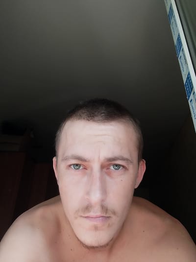 The image shows a close-up, head-and-shoulders selfie of a Caucasian male with short, dark hair and green eyes. He has a light mustache and stubble on his chin. The man is looking directly at the camera with a neutral expression. His shoulders are bare. The background is dark, with a hint of a window frame on the right side of the image. The window frame has a blue and white sticker with text on it. There is also a dark wooden furniture item visible in the lower left portion of the background. The image appears to have been taken indoors. There is no information in the image to suggest a location in the city of Ушья, Russia.