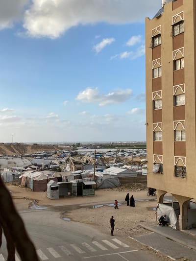 The image depicts a refugee camp in the Sdot Negev Regional Council, Israel. The view is from an elevated perspective, possibly from a building overlooking the camp. The sky is visible, showing a partly cloudy day. The camp consists of makeshift shelters and tents, densely packed together. A road runs alongside the camp, with people walking on it. Some individuals are wearing traditional clothing. A building with multiple windows is visible in the foreground, suggesting the proximity of residential areas to the camp.