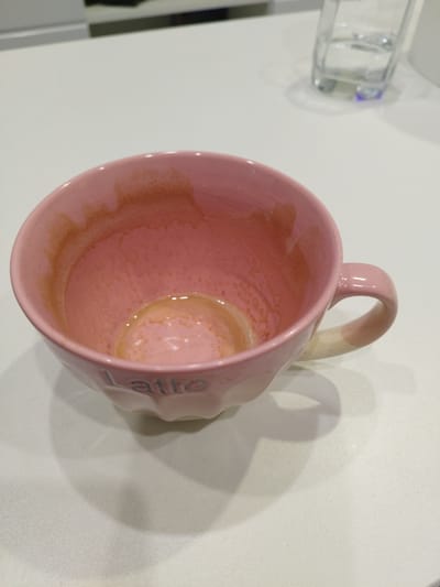 A pink ceramic mug, with the word "Latte" embossed on its exterior, sits on a white countertop. The interior of the mug shows residue from a beverage, with a small amount of liquid remaining at the bottom. The mug has a handle and a slightly flared rim. In the background, a glass of water and part of a kitchen counter are visible. This scene is located on Zonalnaya Stantsiya, Russia.