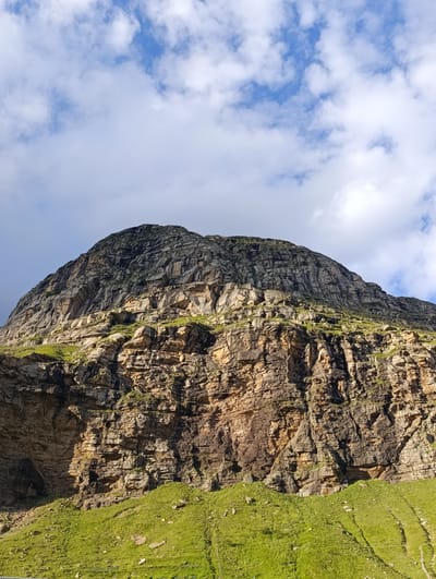 A low-angle, full shot captures a massive, rugged mountain with patches of green grass and vegetation clinging to its lower slopes. The mountain face is composed of layered rock formations, showing variations in color from dark gray and brown to lighter tan hues. Some sections appear sheer and imposing, while others are broken and irregular, with crevices and overhangs. Sunlight illuminates parts of the rock face, creating shadows that emphasize its texture and depth. The sky above is a bright blue, dotted with fluffy white clouds, suggesting fair weather. The location is identified as Damphug, India.