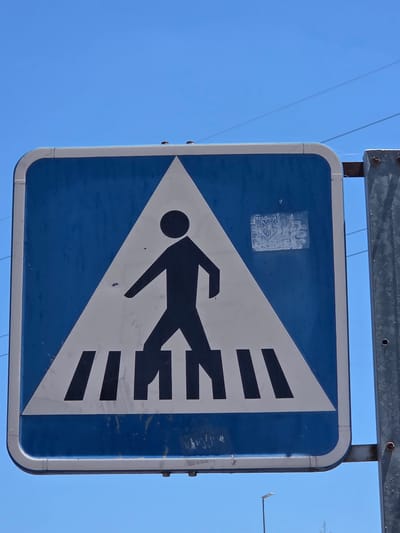 A pedestrian crossing sign is mounted on a metal pole against a clear blue sky. The sign is blue and white, with a white triangle in the center containing a black silhouette of a person walking across black and white stripes. The sign has some wear and tear, with minor scuffs and what appears to be a sticker or graffiti on the blue background. The pole is a silver-colored metal post. In the background, thin wires are visible against the sky, and a street light is faintly visible in the lower right corner. The image appears to be taken from a low angle, looking up at the sign.
