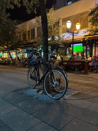 The image depicts a street scene at night in Ilidža, Bosnia and Herzegovina. In the foreground, a black bicycle is parked next to a tree, its front wheel obscuring a metal grate on the sidewalk. The bicycle has a basket on the handlebars. Behind the bicycle, a sidewalk is visible, composed of dark grey paving stones. Further back, a restaurant or cafe area is set up with tables and chairs, occupied by patrons. A large television screen displays a soccer match. The building behind the outdoor seating area features large windows and is illuminated by decorative streetlights. Trees with foliage are interspersed along the street. The overall ambiance is that of a lively urban evening.