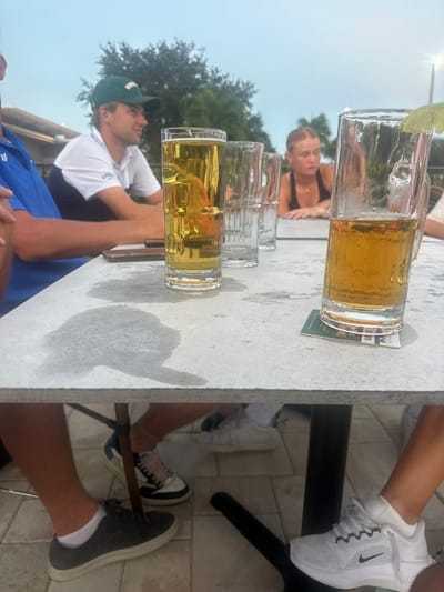 An outdoor patio scene in Estero, United States, depicts four individuals seated around a light grey, rectangular table with a black leg. The foreground features a tall, clear glass containing light amber liquid and ice, directly on the tabletop, displaying condensation. To its right, a shorter, handled glass mug filled with light amber liquid, garnished with a lime wedge, rests on a green and white coaster, also showing condensation. Two empty, textured clear glasses are positioned further back on the table.
On the table's surface, multiple water rings and condensation marks are visible. A dark object, possibly a phone or wallet, is partially obscured near the tall glass.
Three individuals are visible from the waist up. On the left, a person wearing a blue polo shirt has their arm resting on the table. Adjacent, a male wearing a green baseball cap and a white and dark blue polo shirt is seen in profile, looking towards the left. On the right, a blonde-haired female in a dark tank top leans forward with her forearms on the table, looking straight ahead. The fourth person is only indicated by a partial arm on the far right.
Below the table, sections of four pairs of legs and feet are visible on a paved patio surface. These include: a pair of legs with dark grey shoes and white socks; a pair wearing white, black, and pink sneakers; and a pair wearing white Nike sneakers with a black swoosh. The background shows blurred green foliage and an overcast sky, with a faint building structure and a pole light.