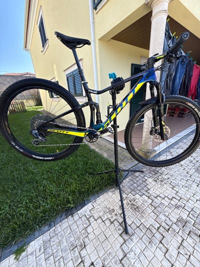 A Scott Spark RC full-suspension mountain bike, featuring a blue-to-black gradient frame with prominent yellow diagonal stripes, is mounted on a black tripod-style repair stand. The bicycle's wheels are elevated above the ground, with "VITTORIA BARZO" visible on the front tire and "VITTORIA MEZCAL" on the rear. The bike exhibits disc brakes, a dropper seatpost, a single-chainring drivetrain, and front and rear suspension components. A blue accessory clamp is attached near the handlebars, and a black water bottle cage is on the downtube.
The scene is outdoors. To the immediate left of the repair stand is a section of vibrant green grass, bordered by a dark curb. To the right and extending under the repair stand, the ground is paved with light-colored, square cobblestones arranged in a diamond pattern. In the background, a two-story building with light yellow stucco walls and a reddish-brown tiled roof is visible. The building features dark-framed windows equipped with louvered shutters. A white, fluted column stands on the right side, supporting an overhang. Behind this column, several items of clothing are suspended on a clothesline. The sky above is clear and blue. The ambient light indicates a sunny day, with shadows cast by the objects. This scene is located in Mealhada, Portugal.