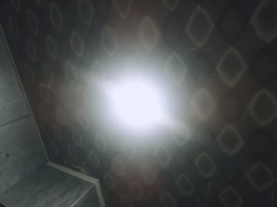 The image shows a corner of a room with patterned wallpaper and tiled walls. A bright light source, likely a lamp or ceiling light, is positioned centrally, causing a strong glare and lens flare effect that obscures some details. The wallpaper has a repeating diamond or leaf-like pattern in dark tones. To the left, a tiled wall is visible, with faint markings that appear to be measurements or labels. The floor or another tiled surface is also partially visible in the lower left. The overall impression is of an indoor space, possibly a bathroom or hallway, under bright artificial lighting. There are no people or specific objects of interest clearly identifiable due to the glare and low light conditions in most of the frame. The location in Aygezard, Armenia, is not discernible from the visual information in the image.