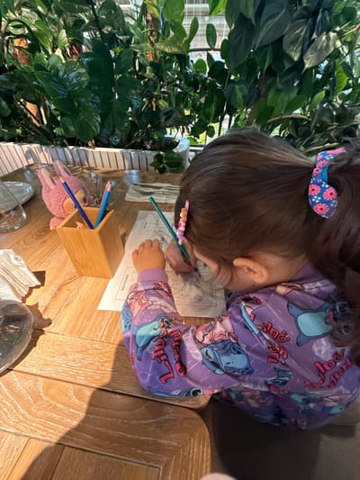 In Агаларов Эстейт Кп, Russia, a young girl with dark hair pulled into a ponytail adorned with a pink and blue floral clip and a smaller beaded clip, is seated at a wooden table, engrossed in coloring a picture. She is wearing a purple long-sleeved shirt featuring a repeating pattern of Stitch characters from Disney's Lilo & Stitch. Her hands are actively engaged with a green crayon, as indicated by its placement and the direction of her grip. In front of her lies a coloring page, and next to it, a small wooden pencil holder contains several colored pencils, including blue and red ones. A plush pink bunny toy is perched on the table near the pencil holder, observing the activity. The table's surface exhibits a prominent wood grain pattern. In the background, lush green foliage is visible, suggesting an indoor setting with abundant plant life, possibly a restaurant or cafe with a natural, biophilic design. A white napkin is placed to the left of the coloring page, and a glass and plate are partially visible on the table.