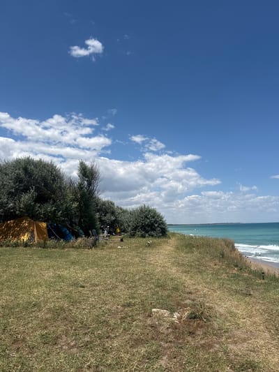 The image depicts a camping scene near the coast in Shabla, Bulgaria. A yellow tent is pitched on a grassy area with several other tents and camping equipment partially obscured by dense green bushes. A table and chairs are set up near the tents, and a few people are visible in the background, one of whom is wearing a yellow dress. The camping area is situated on a clifftop overlooking a turquoise sea with gentle waves rolling onto a sandy beach. The sky is a bright blue with scattered white clouds, suggesting a clear and sunny day. The environment is natural and rugged, with dry grass and wild vegetation dominating the foreground and the edge of the cliff.