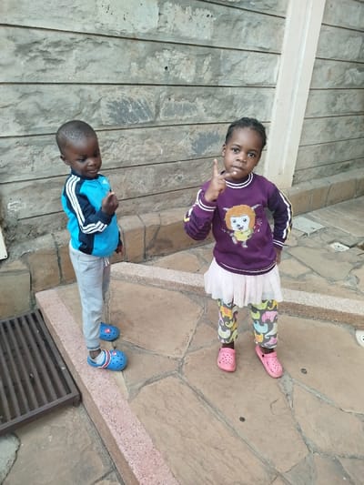 Two young children stand outside. The child on the left wears a blue and black jacket, gray pants, and blue Crocs. He holds his fingers together. The child on the right wears a purple sweater with a graphic, a white skirt, patterned leggings, and pink Crocs. She holds up a finger. They stand on tan stone pavers in front of a stone wall with a white trim piece to the right. A metal grate is to the left.