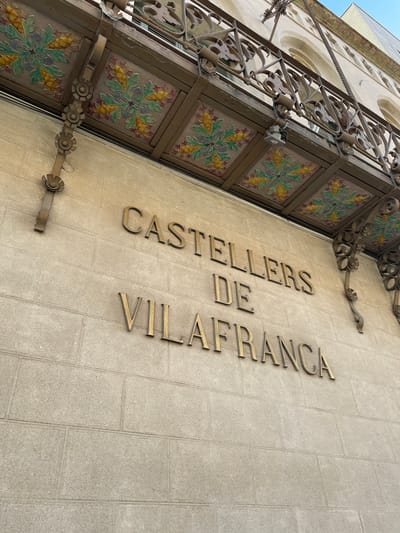 The image shows the facade of a building with the name "CASTELLERS DE VILAFRANCA" in large, golden, three-dimensional letters. The building's facade is made of light-colored stone blocks. Above the lettering, there is an ornate balcony with decorative tiles and wrought iron railings. The balcony has a metal structure supporting it, adorned with decorative floral patterns in vibrant colors, including green, yellow, blue, and red. The overall aesthetic suggests a historical or traditional building, likely a cultural center or the headquarters of a local group, given the name "Castellers," which refers to human tower builders. The scene is captured from a low-angle perspective, looking upwards towards the building. The sky is visible at the top right corner of the image, indicating an outdoor setting during the daytime. The architecture and decorative elements are characteristic of Catalan modernist or noucentista styles, commonly found in towns like Vilafranca del Penedès, Spain, which is renowned for its human tower traditions.
