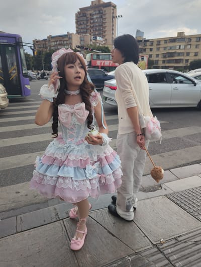 In Xishan District, China, two individuals are standing on a pedestrian crossing. The person on the left is a woman wearing a pink and blue Lolita-style dress with lace trim, a choker, and a matching headpiece. She is holding a mobile phone to her ear and has a small box in her other hand. The person on the right is a woman wearing a cream-colored cardigan over white pants and black shoes. She is carrying a fluffy white purse with pink accents and holding a wooden stick with a brown pom-pom at the end. The background shows a street scene with a purple bus, a red bus, several cars, and buildings in the distance. The sky is overcast.