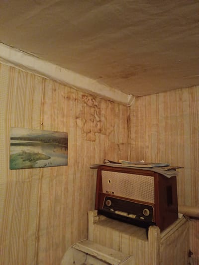 The image depicts an interior scene in what appears to be a dilapidated room. The wallpaper has vertical stripes, with water damage and stains visible on the upper parts of the walls, particularly near the corner of the room. A framed landscape painting hangs on the wall. An old, wooden radio sits atop a small, possibly makeshift, stand or stack of objects. Several sheets of paper are scattered on top of the radio. The ceiling has a similar stained and worn appearance. The bottom of a white metal object is also present. The location is stated as Verkhnii Dvor, Russia.