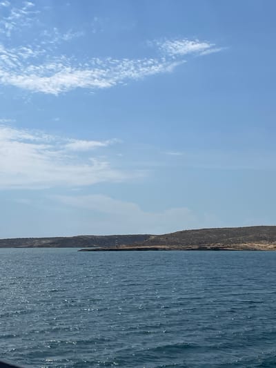 A landscape photograph depicting a body of water, likely a sea or ocean, under a blue sky with scattered clouds. On the right side of the frame, a strip of land, possibly a cliff or hillside, runs vertically. In the center of this land strip, there is a structure resembling a lighthouse. The water surface exhibits texture indicative of wavelets or ripples. The scene is illuminated by daylight. The location is identified as Unknown, Venezuela.