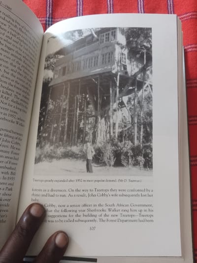 The image is a black and white photograph of a large treehouse in a forest. The treehouse has two levels, a roof, and multiple windows. The treehouse is built on a thick tree trunk and supported by wooden beams. There are two people standing near the base of the treehouse, one facing the camera and the other facing away. The image is likely from the city of Ganja, Azerbaijan, based on the provided coordinates.  The image has a caption beneath it that reads, "Treetops greatly expanded after 1952 to meet popular demand. (Mr. D. Steerman)".  The text further states that on the way to Treetops, they were confronted by a rhino and had to run. As a result, John Cobby’s wife subsequently lost her baby.  Cobby, now a senior officer in the South African Government, that the following year Sherbrooke Walker rang him up in his suggestions for the building of the new Treetops-Treetops it was to be called subsequently. The Forest Department had been 
107.