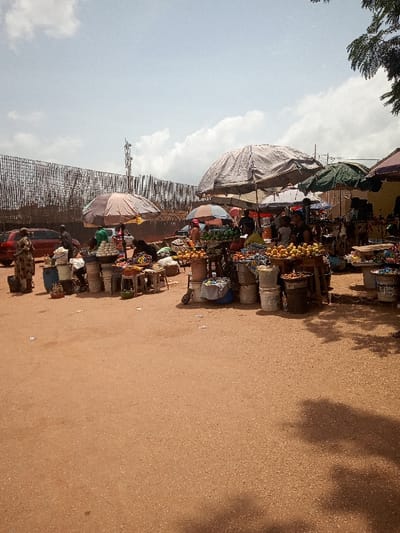 The image depicts a street market in Otta, Nigeria, located at coordinates 6.682 latitude and 3.168 longitude. The market is set up on a dirt road, lined with various stalls, each covered by a canopy or an umbrella.  There are several people at the stalls, some of whom are selling produce like fruits and vegetables, while others are shopping. There are also buckets and other containers on the ground, used for storing and transporting items. The scene is captured under a partly cloudy sky.  A tall communication tower can be seen in the background.  Several cars are parked on the road, behind the market stalls.  A wooden fence with a bamboo mat-like structure is positioned behind the cars.