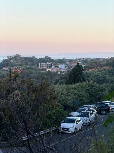 The image shows an elevated view of Acireale, Italy, likely taken from a hillside. The sky is light with a soft pinkish hue suggestive of dawn or dusk. In the middle ground, there are buildings and vegetation, including trees. On the right side of the image, a road is visible with several parked cars. The foreground includes branches of a tree.