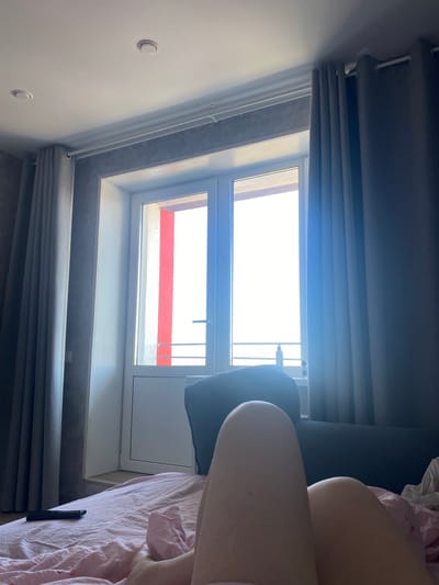 The image, oriented sideways, depicts an interior room, likely a bedroom, illuminated by natural light. In the foreground on the right, a person is partially visible, showing their legs and lower torso resting on a bed covered with a pink and white striped duvet. A dark grey pillow is also partly seen near the person.
The central feature is a large, white-framed window, possibly a skylight or dormer window, through which bright daylight streams into the room. The window appears to have two panes with a visible handle on the upper section. Outside the window, a clear, sunlit view reveals a structure with distinct horizontal red and white stripes, suggesting a sunny daytime. Below the window, dark grey pleated blinds or roller blinds are partially drawn. The interior walls are plain white, and two circular white light switches or fixtures are mounted on the left wall. The bright lighting conveys a serene and relaxed atmosphere. No text is visible in the image. The location context for this scene is Zubovo, Russia.